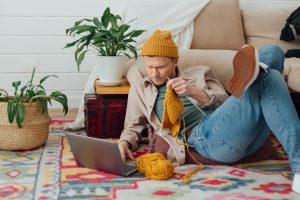 Young serious man looking at laptop. Man learning new hobby, knitting on needles. Knitting project in progress. - Image